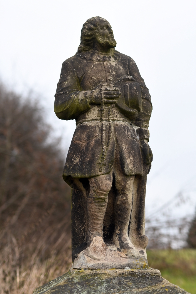 A stone figure of a man holding a rosary and hat, standing on a plinth. The figure has a beard and is dressed in traditional costume. In the background are blurred trees and sky.