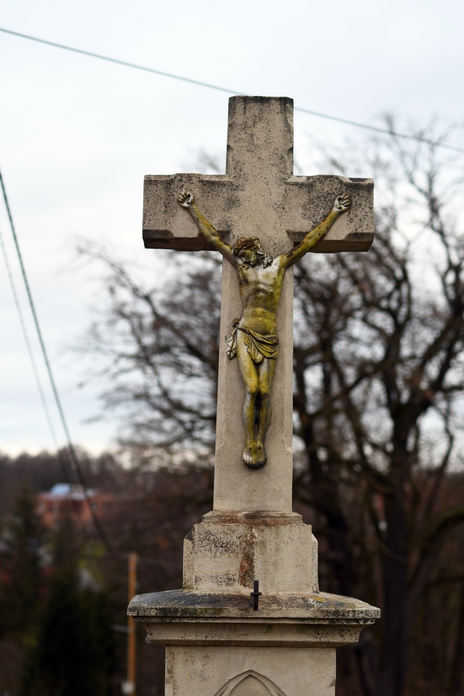 A stone cross with a faded figure of Christ against a background of bare trees and distant buildings on the Czech side of Těšín Silesia.
