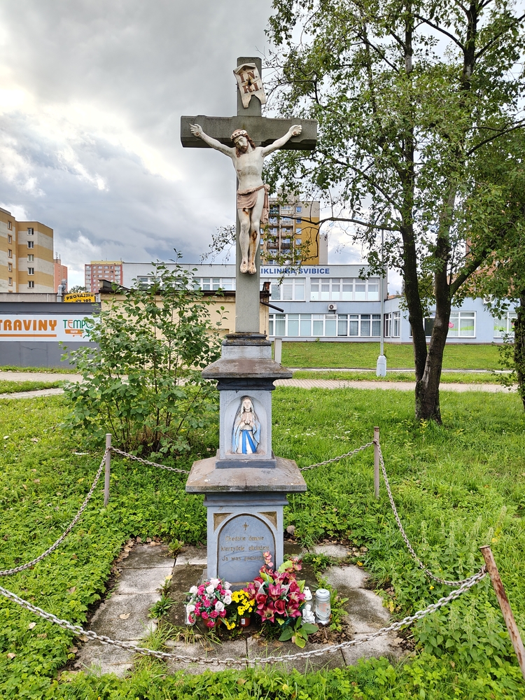 Roadside cross with a statue of Christ in front of a modern building. On the plinth is a statue of the Virgin Mary and an inscription. Flowers and a candle are placed at the base, surrounded by a chain-link fence.