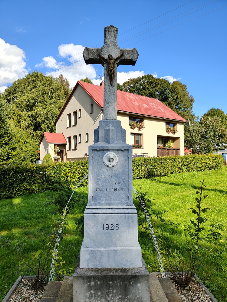 A stone cross with a statue of Christ stands on a plinth with the inscription 'Jesus Christ, have mercy on us' and the date 1928. In the background is a house with a red roof.