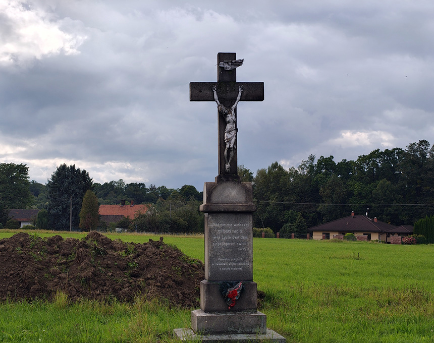 A stone crucifix stands in a grassy field with a cloudy sky. The cross features a figure of Christ and an inscription on the base. In the background, there are trees and a few houses.