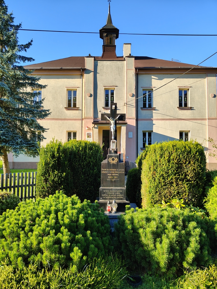 Пам'ятник Першій світовій війні перед бежевою будівлею з маленькою вежею, оточений зеленими кущами. Розп'яття стоїть на чорному постаменті з написами та свічками біля основи.