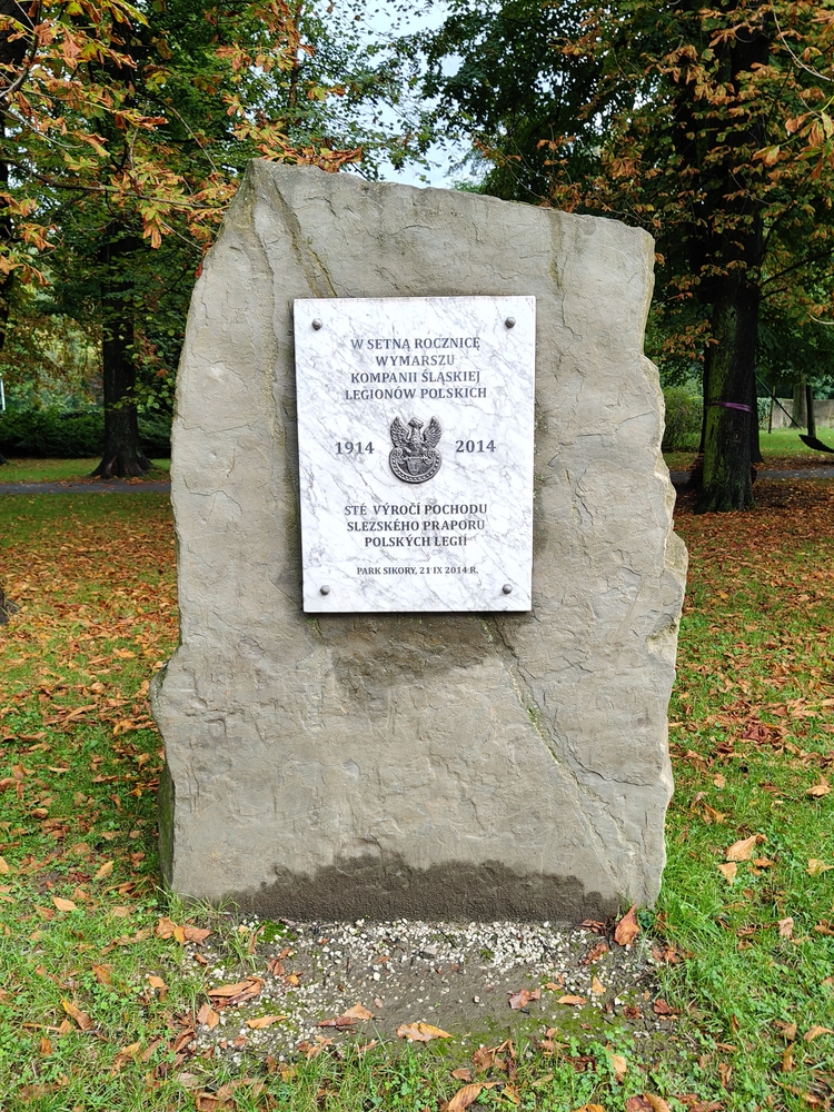 Кам'яний пам'ятник з меморіальною дошкою на честь 100-річчя походу Сілезької роти польських легіонів, розташований у парку з деревами і травою.