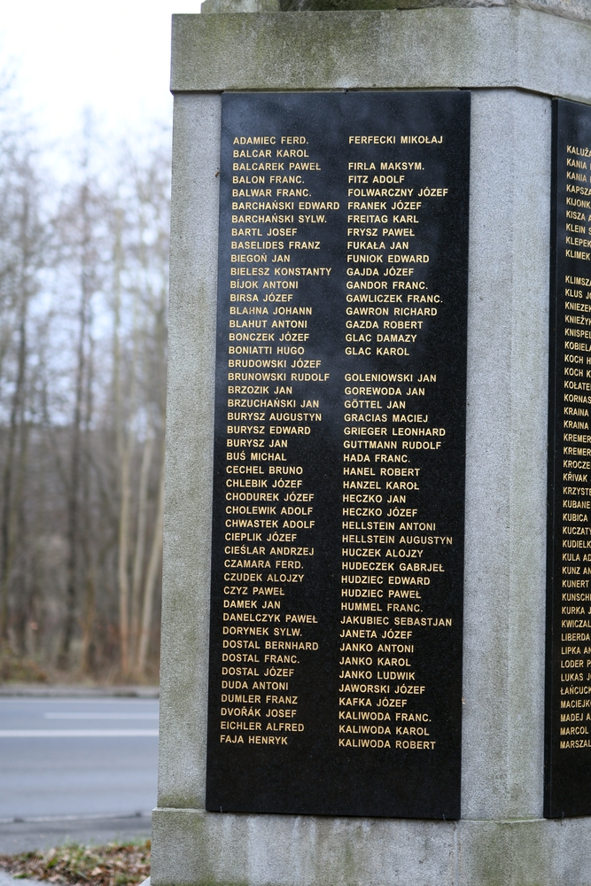 Пам'ятник у Карвіні, що вшановує жертв Першої світової війни, з кам'яною структурою та вигравіруваними іменами загиблих солдатів. На задньому плані видно дерева та дорогу.