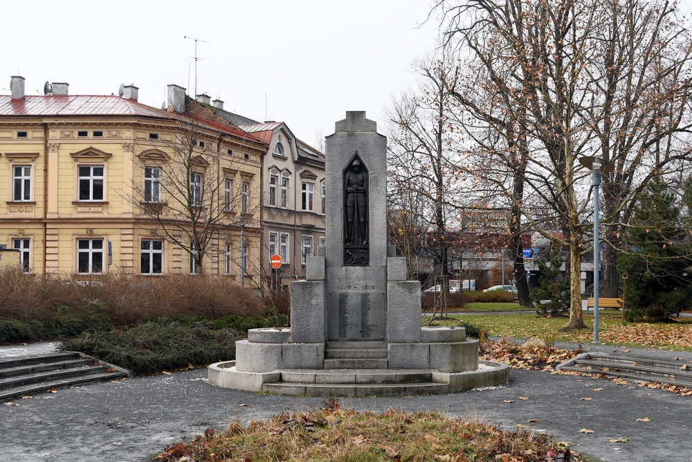 Пам'ятник Першій світовій війні в Тешинській Сілезії, із солдатом у ніші на гранітній стелі. Навколишній парк з лавками та деревами, поруч із історичними будівлями.