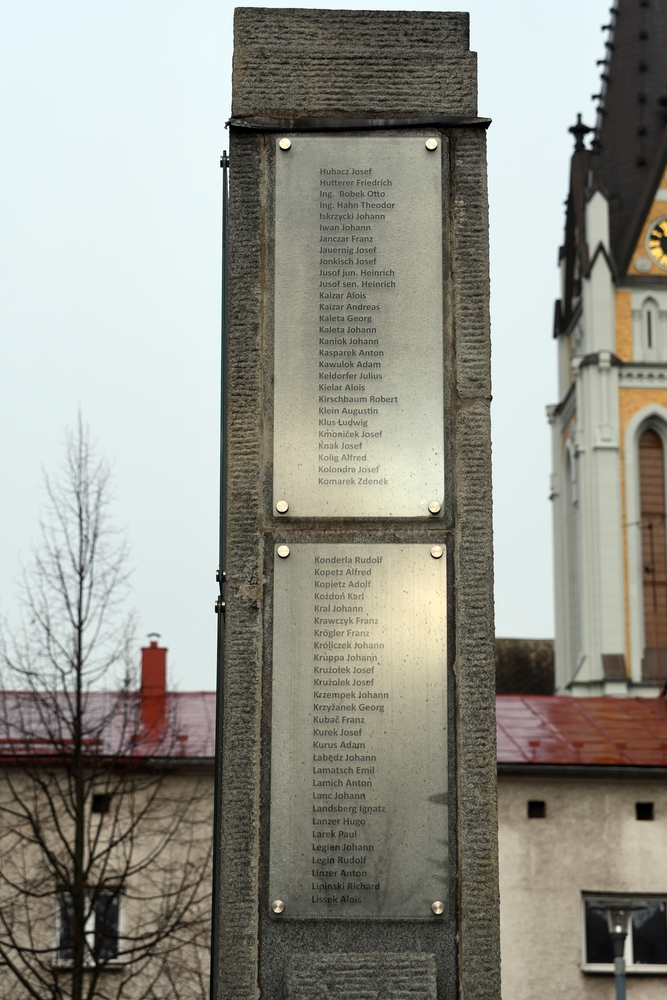 Кам'яний пам'ятник з двома металевими табличками з іменами жертв Першої світової війни. На задньому плані видно церкву з годинниковою вежею.