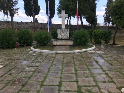 Marmurowy pomnik z krzyżem i płaskorzeźbą orła na grobie zbiorowym polskich żołnierzy w Missolungi, Grecja. Otoczony zielenią i flagami Grecji oraz Polski.