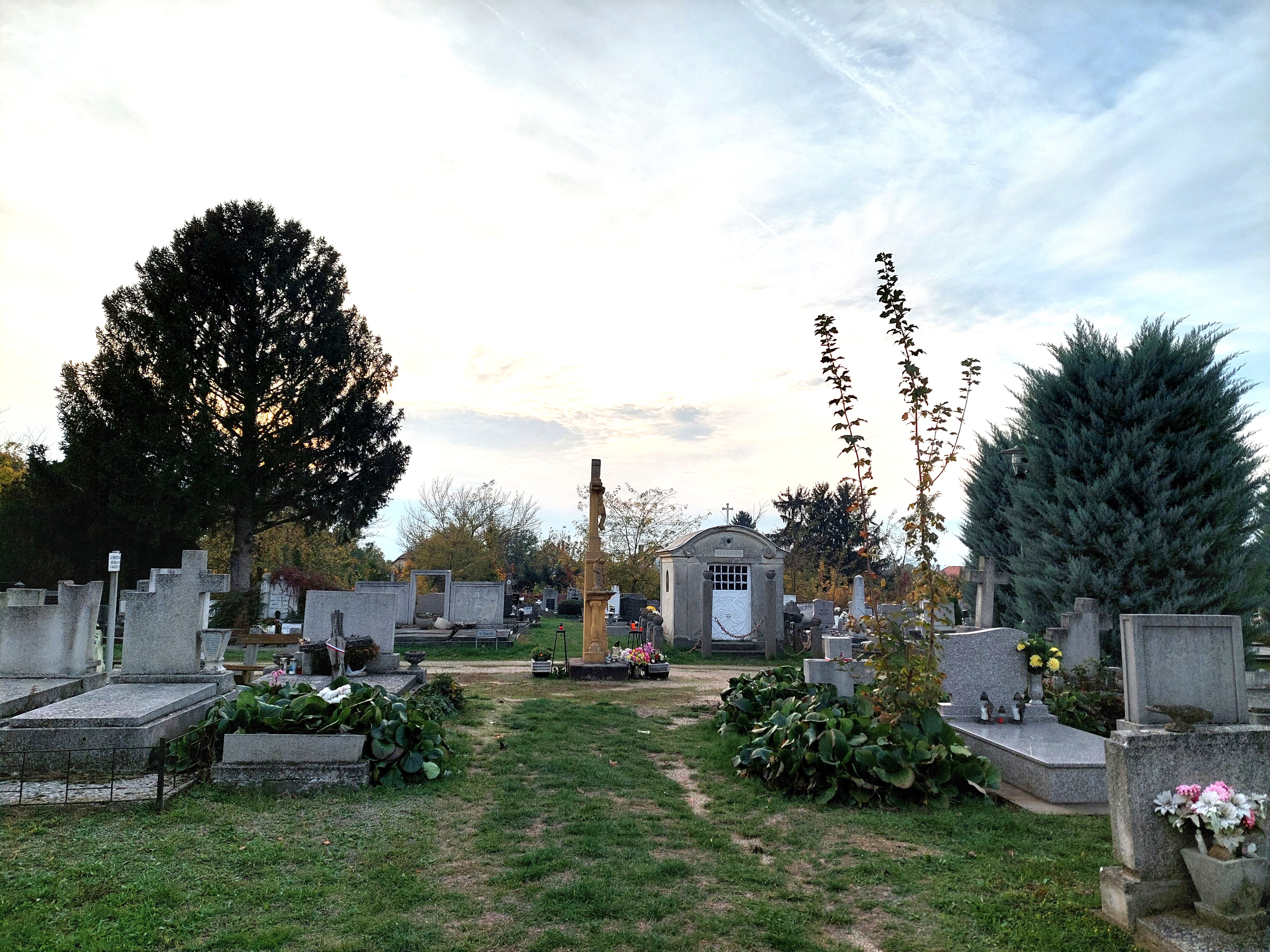 Catholic cemetery with four Polish refugee graves. Graves are near a large cross with a figure of Jesus, along an alley separating plots III and IV. Visible are stone graves, plants, and a mausoleum in the background.
