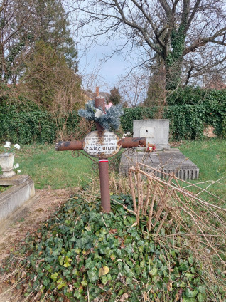 Grób polskiego żołnierza Józefa Zająca z żelaznym krzyżem i metalową tabliczką z inskrypcją w języku polskim i węgierskim, otoczony bluszczem i suchymi gałęziami, na cmentarzu z drzewami i kamiennym pomnikiem w tle.