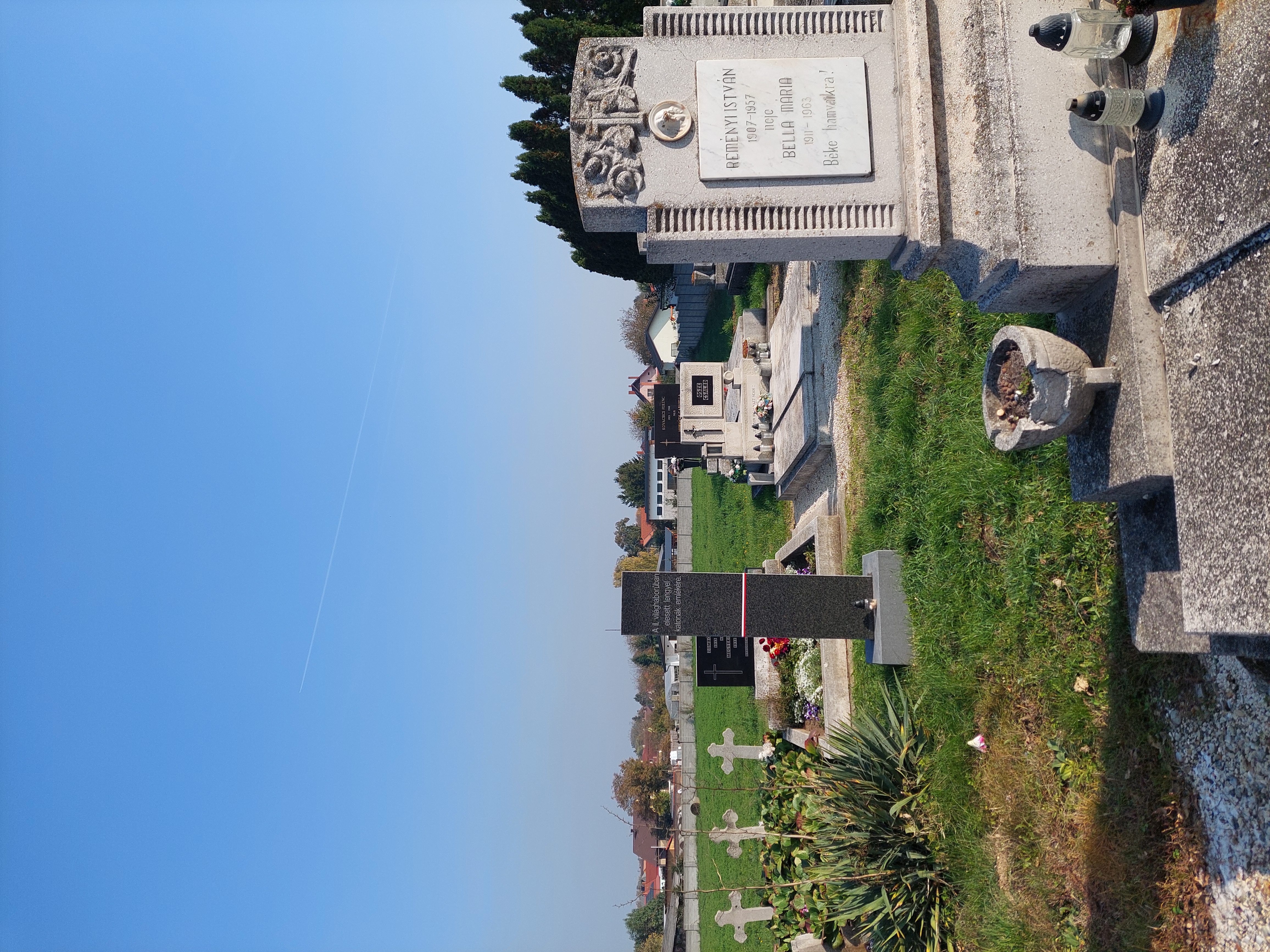 Graves of Polish soldiers in Fertőszentmiklós cemetery. A dark granite obelisk with Hungarian inscription stands among other tombstones. Green grass and a clear blue sky in the background.
