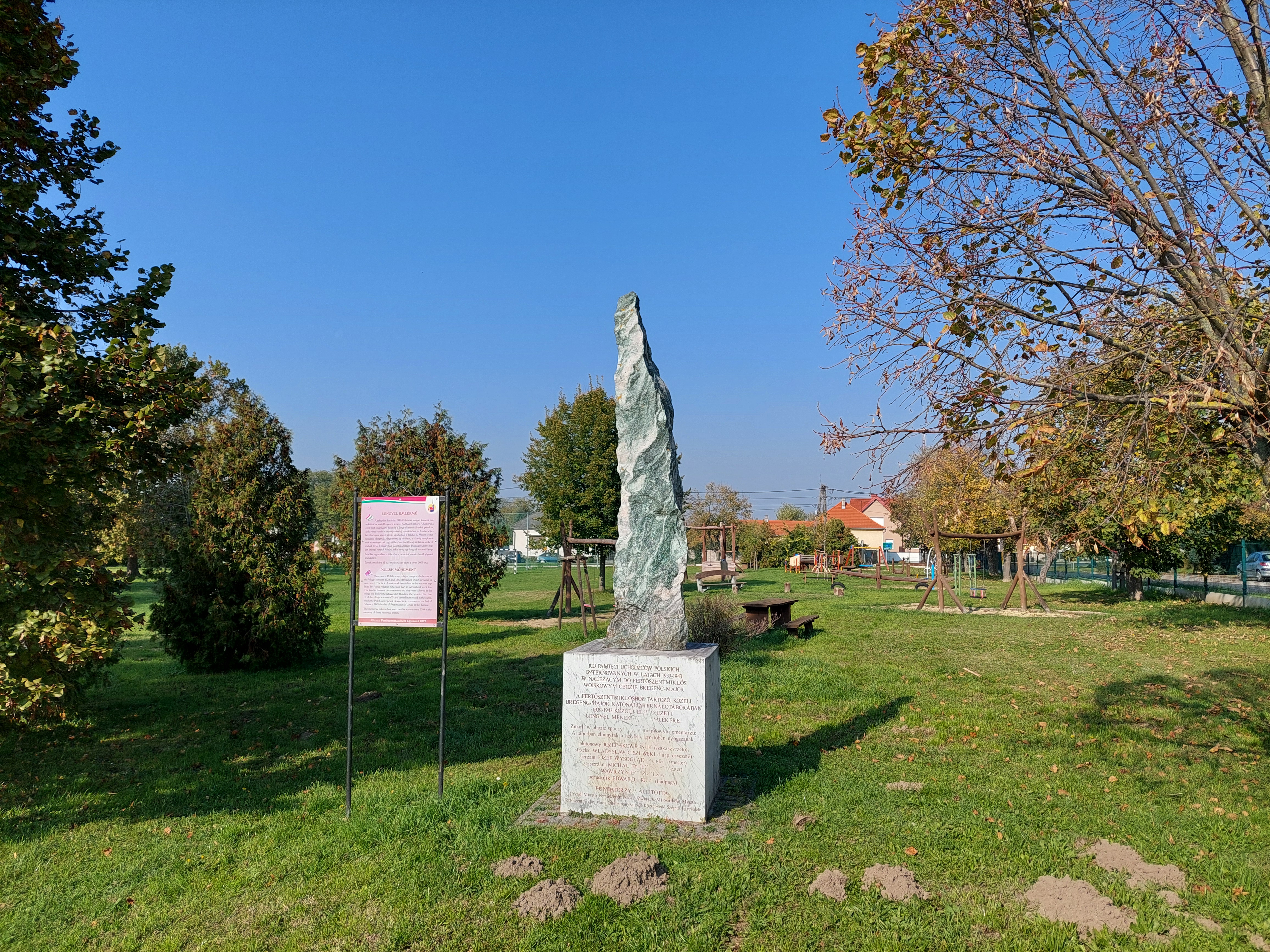 A granite obelisk commemorating Polish soldiers from World War II stands in a grassy area with trees and a playground in the background. A sign with text is placed next to the monument.