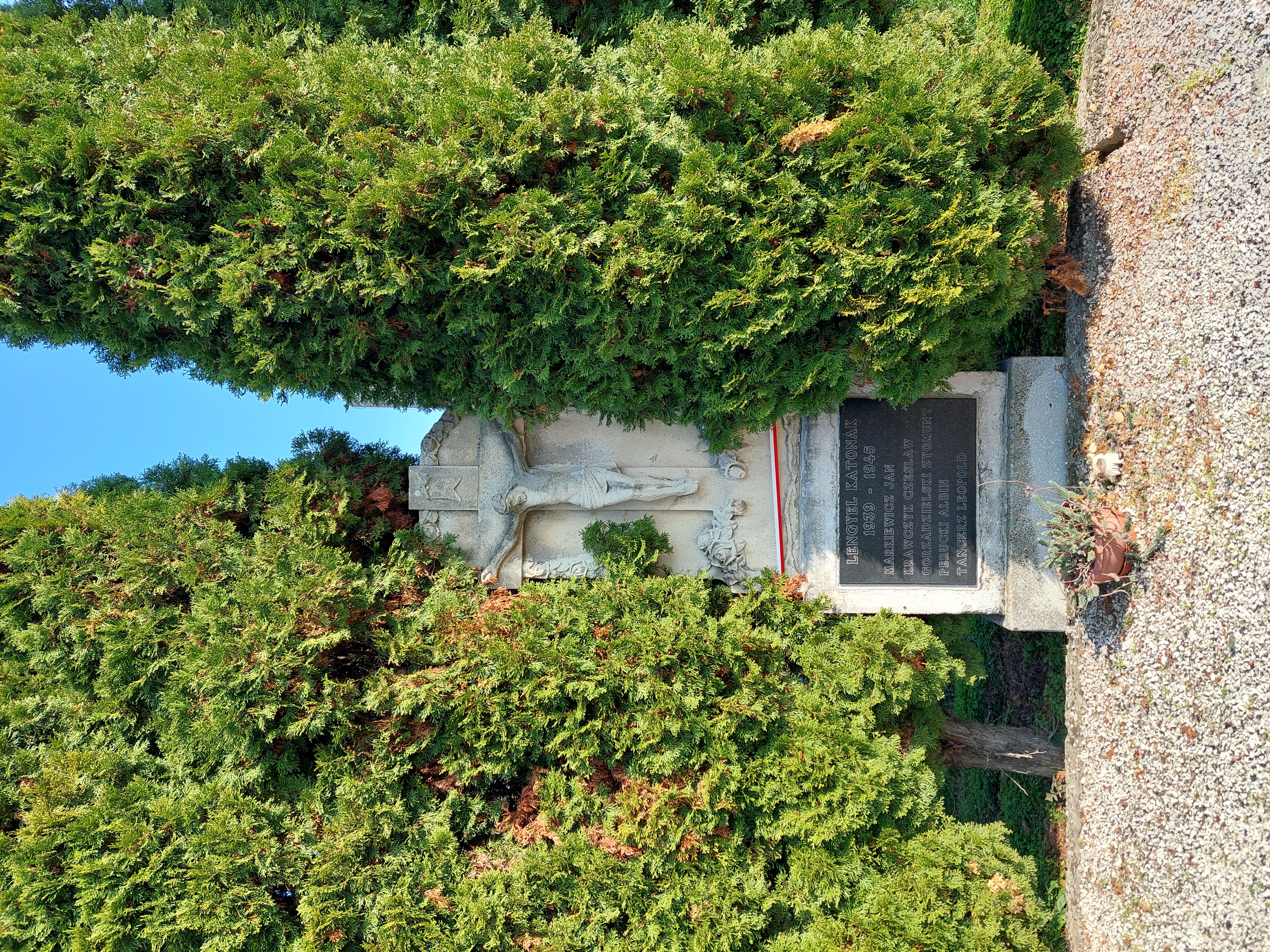 A stone monument with a large crucifix and a dark plaque inscribed with names: Jan Markiewicz, Czesław Krawczyk, Zygmunt Gorzadzielski, Albin Perucki, Leopold Tancerz, surrounded by tall trees.