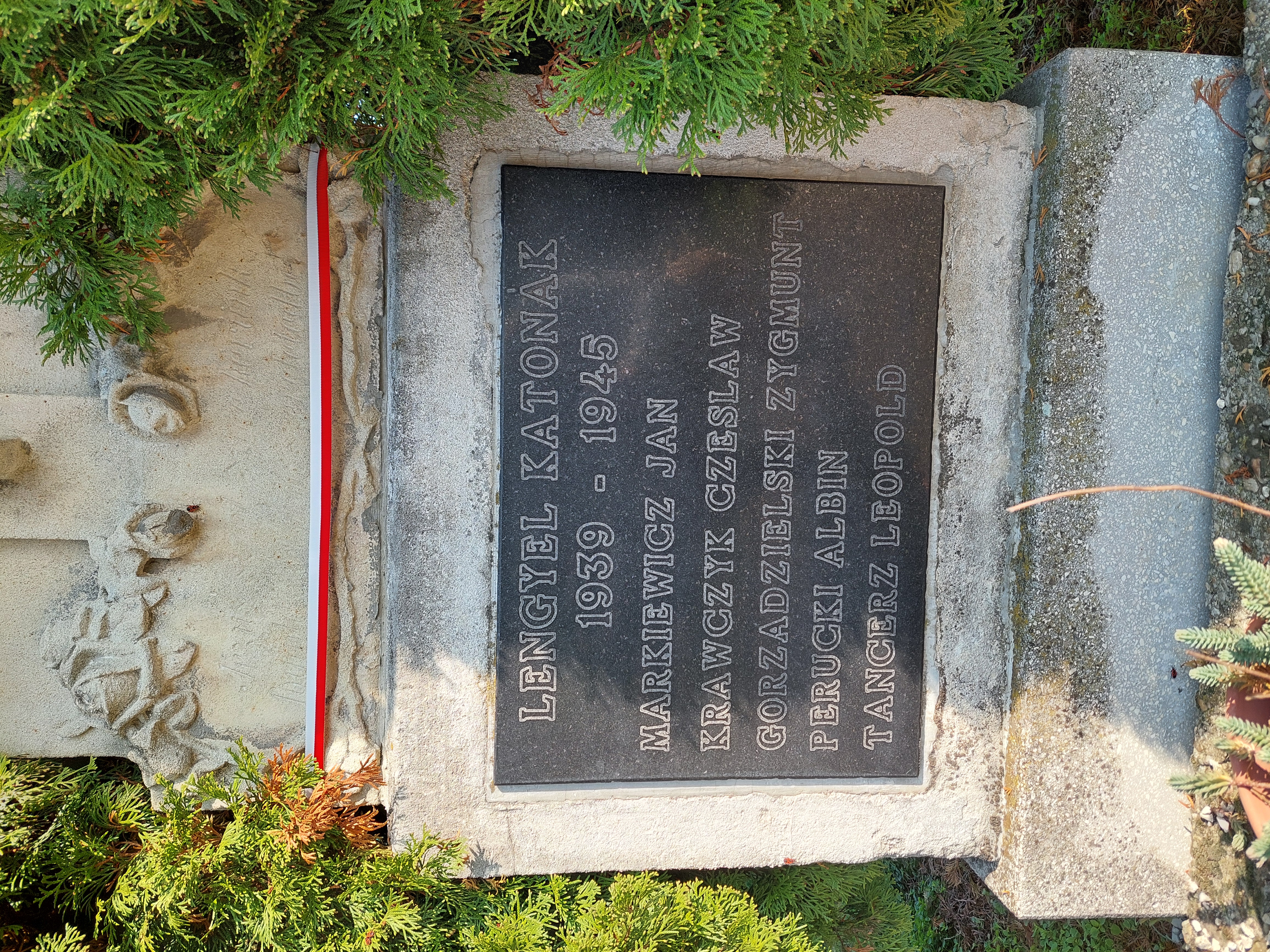 Gravestone with a dark plaque inscribed with names: Markiewicz Jan, Krawczyk Czeslaw, Gorzadzielski Zygmunt, Perucki Albin, Tancerz Leopold, and dates 1939-1945. Surrounded by greenery.