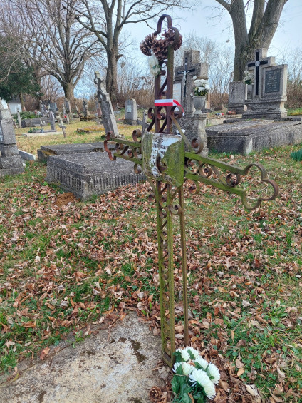 Grób polskiego żołnierza Adama Turczyńskiego w Kislőd na Węgrzech. Metalowy krzyż z uszkodzoną tabliczką stoi na betonowej płycie otoczonej opadłymi liśćmi. U podstawy leżą białe kwiaty.