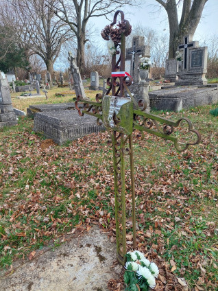 Grób polskiego żołnierza Adama Turczyńskiego w Kislőd na Węgrzech. Metalowy krzyż z uszkodzoną tabliczką stoi na betonowej płycie otoczonej opadłymi liśćmi. U podstawy leżą białe kwiaty.