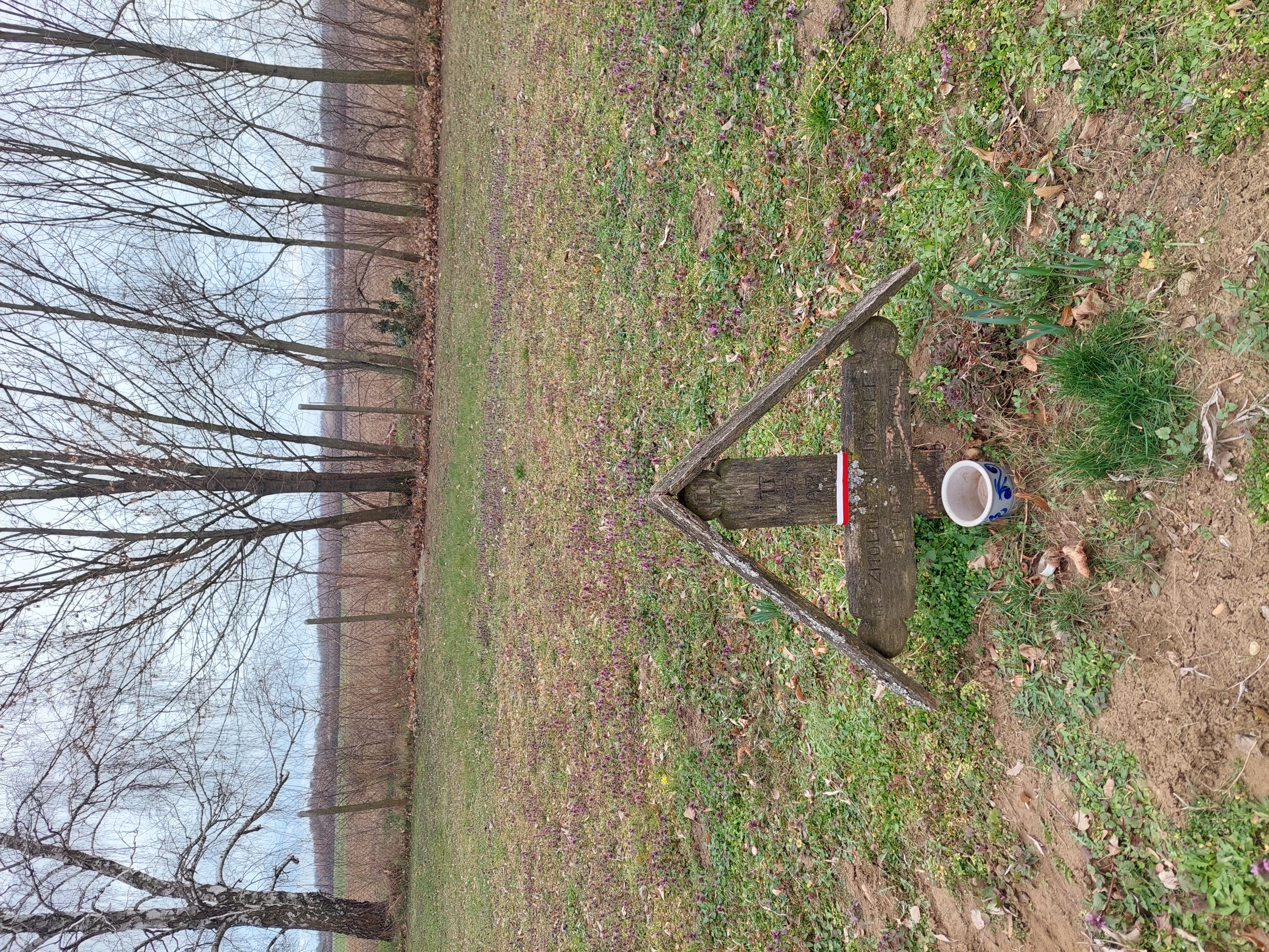 Croix en bois marquant la tombe du réfugié polonais Józef Smoliński à Oroszi, Hongrie. La croix porte une inscription hongroise partiellement effacée et un petit pot en céramique à sa base. Des arbres et un champ en arrière-plan.