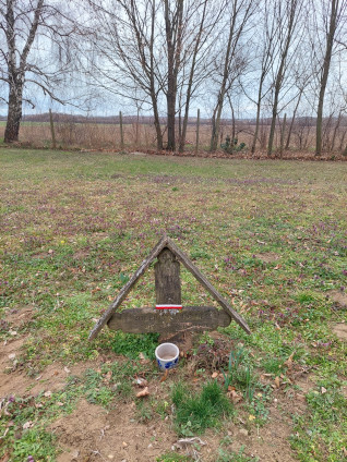 Drewniany krzyż oznaczający grób polskiego uchodźcy Józefa Smolińskiego w Oroszi na Węgrzech. Krzyż ma częściowo zatarty napis węgierski i mały ceramiczny pojemnik u podstawy. W tle drzewa i pole.