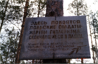 Metalowa tablica na drzewie z rosyjskim tekstem upamiętniającym polskich żołnierzy zmarłych w niewoli. W tle las z wysokimi drzewami.