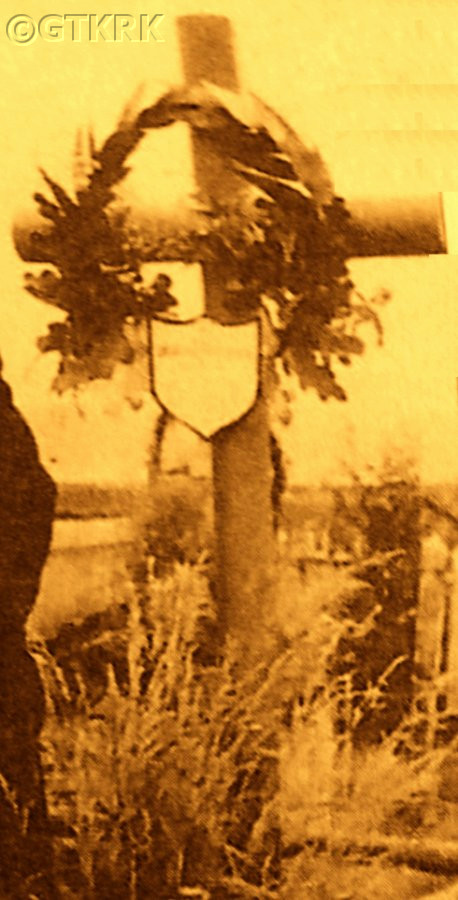 A sepia-toned photograph of a grave marked by a wooden cross adorned with a wreath. A plaque with Polish text is attached to the cross. Grass and a fence are visible in the background.