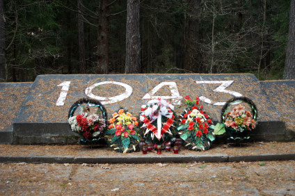 Kamienny pomnik z wyrytym rokiem 1943, otoczony wieńcami i kwiatami, w leśnym otoczeniu.