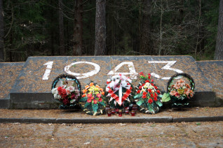 Kamienny pomnik z wyrytym rokiem 1943, otoczony wieńcami i kwiatami, w leśnym otoczeniu.