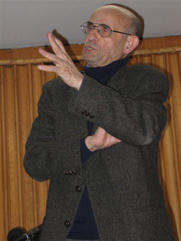 A man in a dark suit and glasses gestures with his hand, standing in front of a wooden wall.