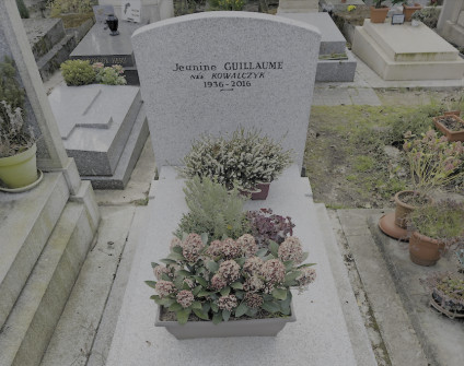 Nagrobek Joanny Kowalczyk z ćwierćłukowym zwieńczeniem, z inskrypcjami, otoczony doniczkowymi roślinami i kwiatami.