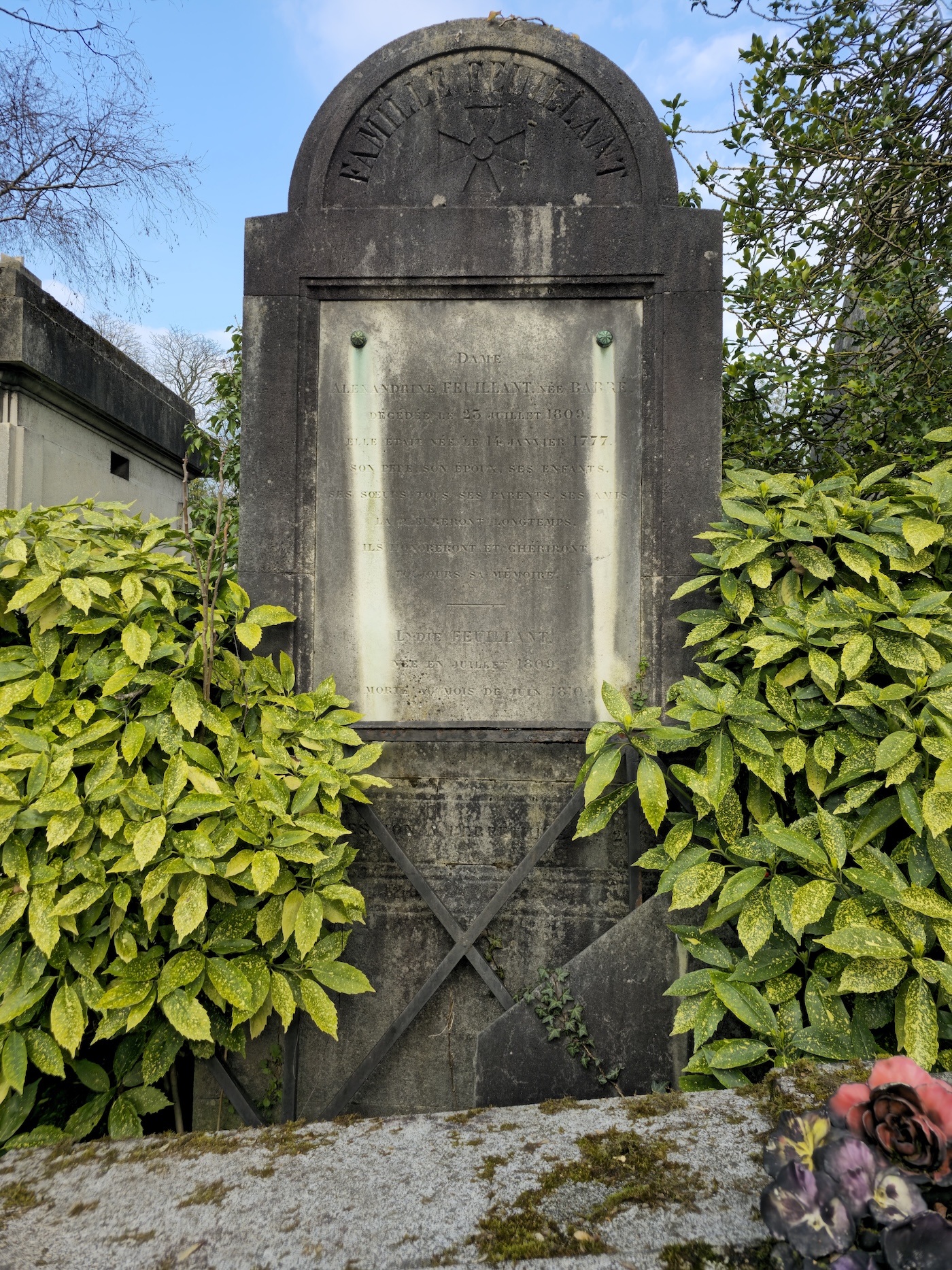 Pierre tombale de la famille Feuillant avec un relief de croix sur le tympan. Entourée de verdure, la pierre porte des inscriptions. Un bouquet de fleurs artificielles est au premier plan.