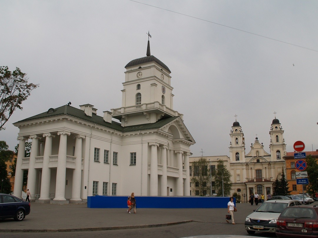 Римсько-католицький собор у Мінську з бароковими вежами та прилеглою неокласичною будівлею. На передньому плані видно автомобілі та пішоходів.