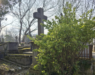 Wysoki krzyż na postumencie z inskrypcjami, otoczony zielenią i innymi grobami. Scena rozgrywa się na cmentarzu z nagimi drzewami w tle.