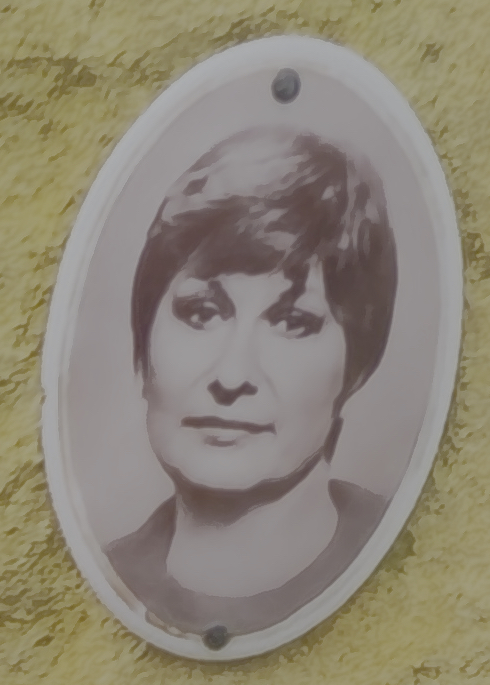 Plaque ovale en céramique avec un portrait en noir et blanc d'une femme aux cheveux courts, fixée sur un mur jaune.