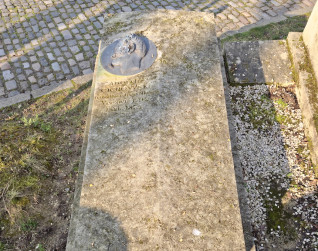Nagrobek Adolfa Cichowskiego z brązowym medalionem po lewej stronie. Inskrypcja poniżej: 'Adolf Cichowski, Warszawa 1794, Paryż 1854'. Kamień jest zwietrzały z mchem.