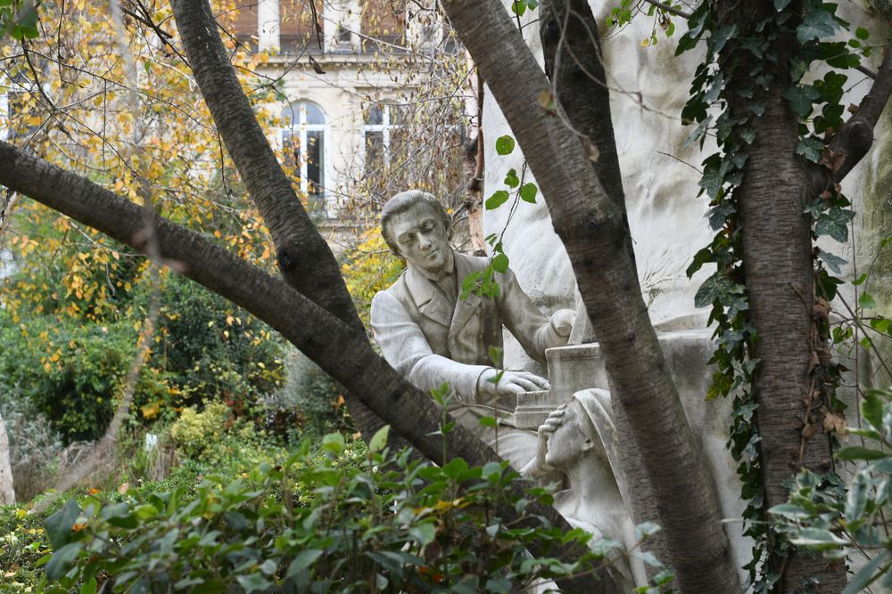Statue of Frédéric Chopin in Monceau Park, Paris, partially obscured by trees. Chopin is depicted playing the piano, with a woman symbolizing Hearing at his feet.