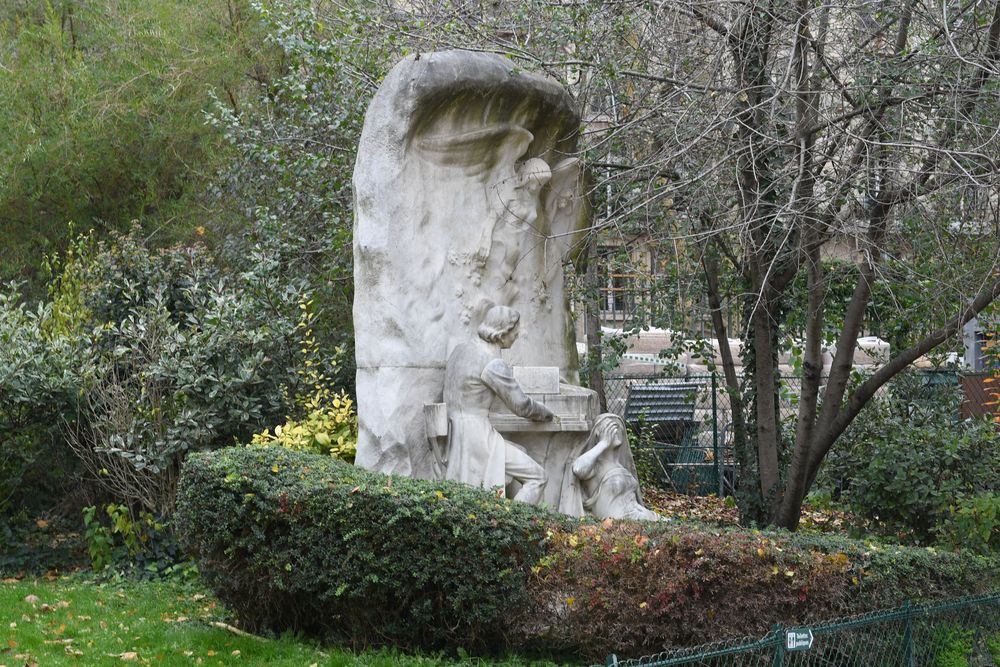 Monument of Frédéric Chopin in Monceau Park, Paris, depicting Chopin seated at a piano with a winged Muse above and a woman symbolising Hearing at his feet, surrounded by greenery.
