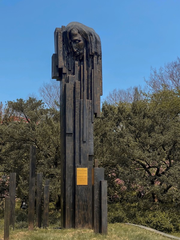 Statue en bronze de Frédéric Chopin dans le parc Zhongshan de Shanghai, avec des colonnes verticales abstraites ressemblant à des touches de piano et au profil émergent de Chopin. À l'arrière-plan, des arbres et un ciel clair.