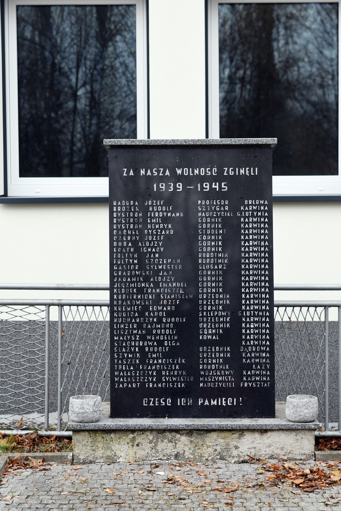 Пам'ятник у Петровицях біля Карвіни, що вшановує жертв Другої світової війни, з чорною табличкою з іменами та професіями загиблих, на тлі будівлі з двома вікнами.