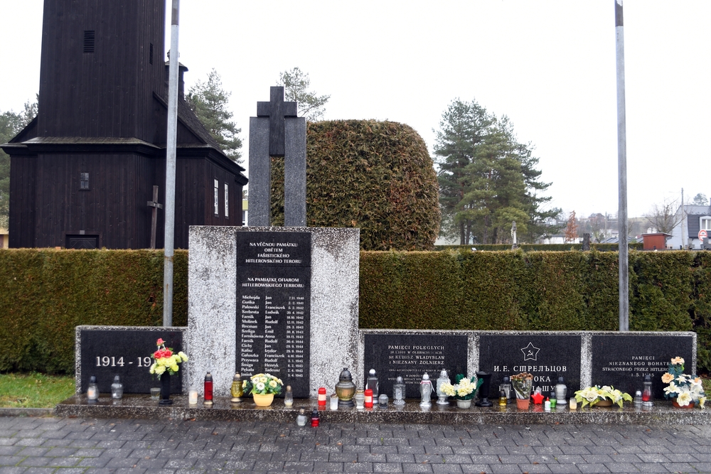 Пам'ятник у Петрівцях біля Карвіни, що вшановує жертв Другої світової війни. Гранітні плити з написами та іменами, оточені свічками та квітами. На задньому плані дерев'яна будівля та дерева.