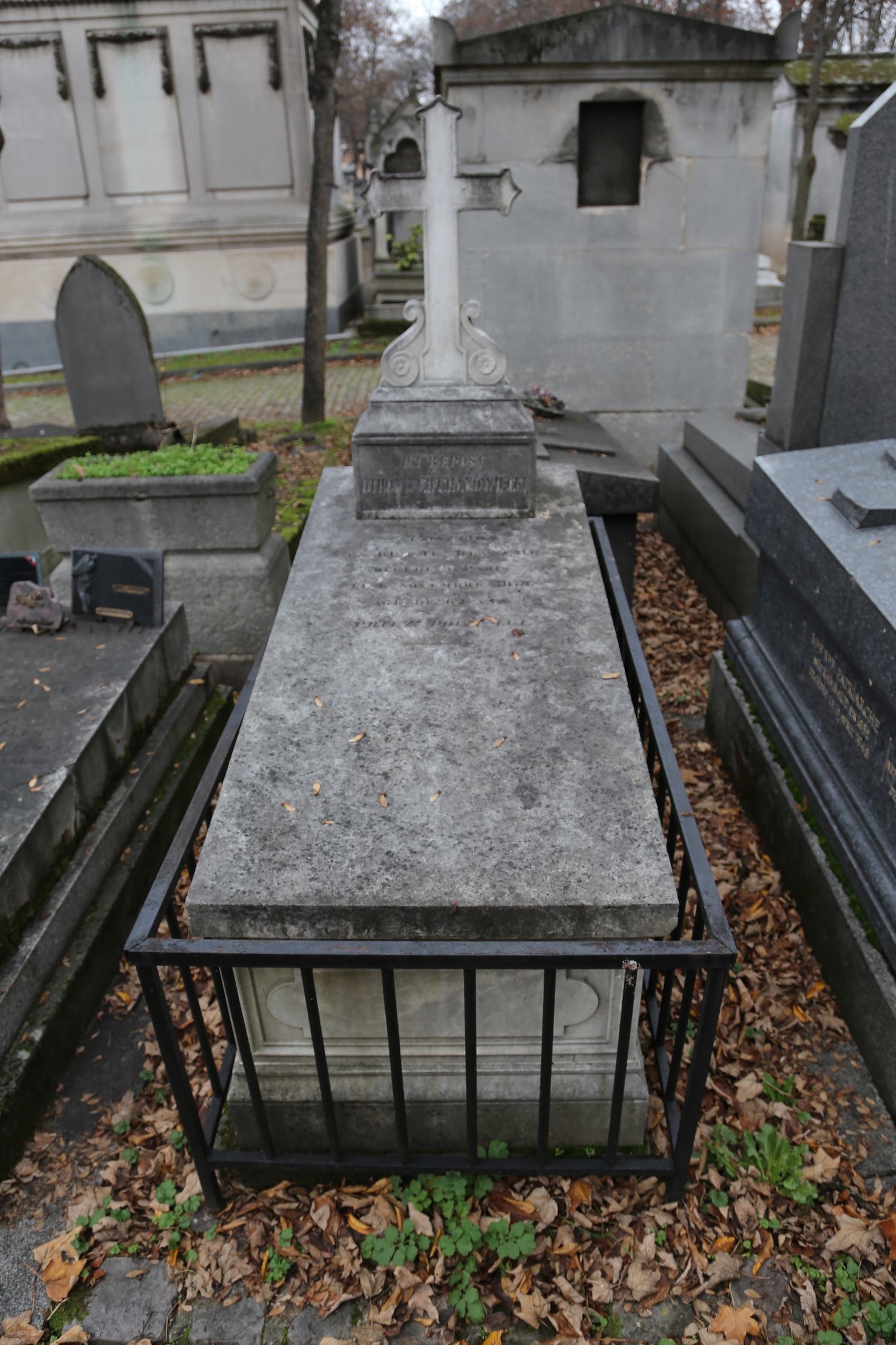 Tombe d'Otto Ciechanowiecki avec une croix sur une base rectangulaire, entourée d'une clôture en fer. Des feuilles mortes couvrent le sol autour de la tombe.