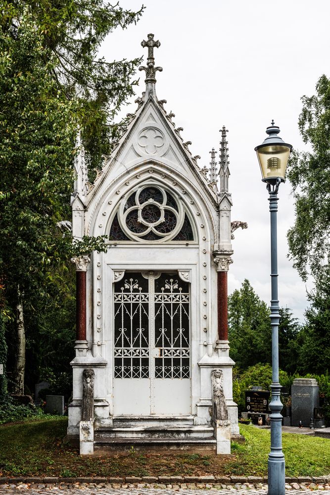 The facade of the Grauer family chapel in Opava, with its neo-Gothic design, sharp arches and decorative pinnacles. A lantern stands on the right, surrounded by trees and gravestones.