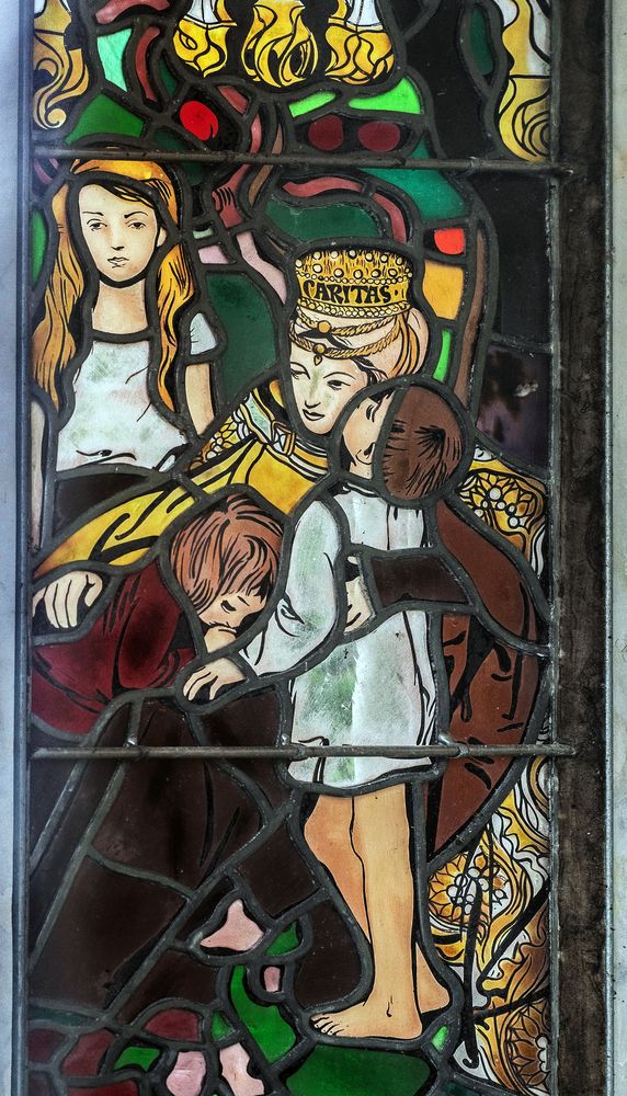 A stained glass window by Jozef Mehoffer in the Grauer family chapel in Opava, depicting a woman wearing a crown with the inscription 'Caritas' embracing two children. The scene is surrounded by vivid colours and intricate patterns.