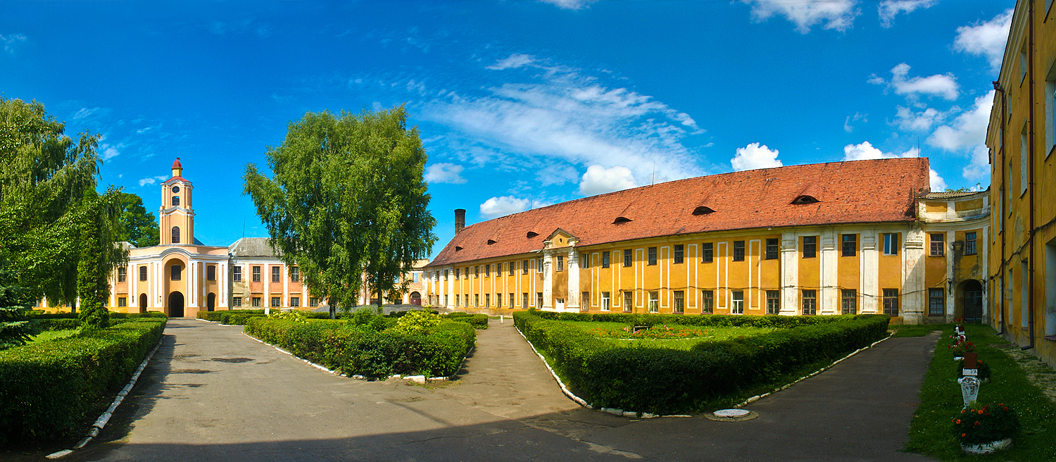 Pałac Radziwiłłów w Ołyce ma żółtą fasadę z wieżą zegarową przy wejściu. Budowla tworzy czworobok z dziedzińcem, otoczona zielenią i ścieżkami.