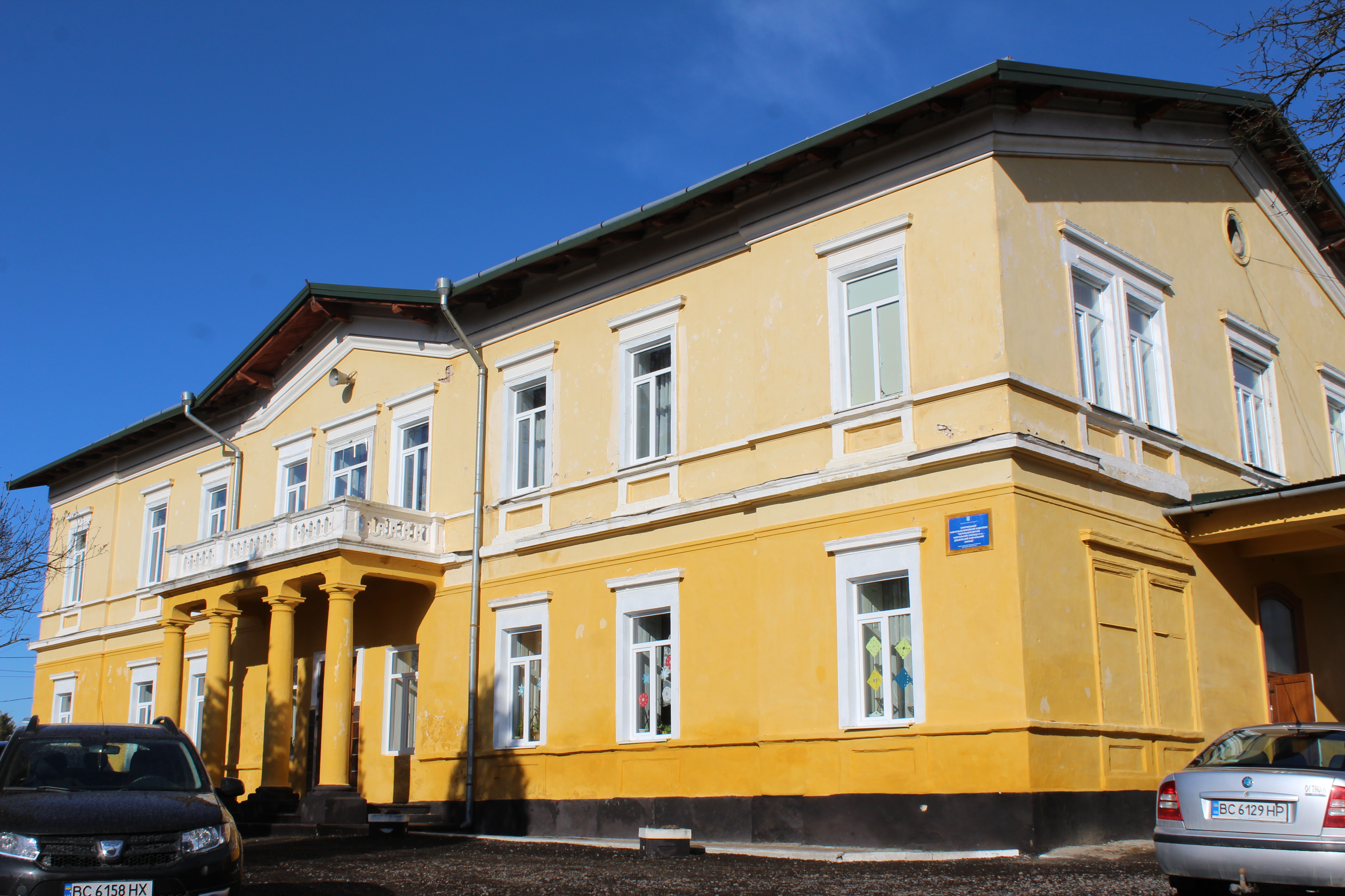 Жовтий цегляний маєток у Снятинці з білими рамами вікон і невеликим балконом на колонах. Перед будівлею припарковані дві машини. Будівля під ясним небом.