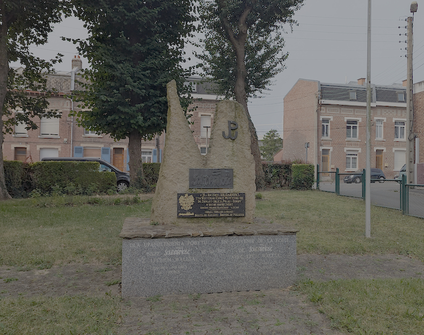 Monument à 'Solidarité' à Lens, situé près de l'église du Millénaire. Présente une structure en pierre avec le logo 'Solidarité' et des plaques commémoratives. Arbres et bâtiments résidentiels en arrière-plan.