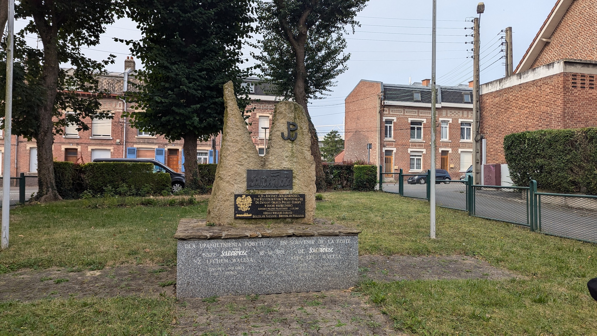 Monument à la 'Solidarité' à Lens, situé près de l'église du Millénaire. Érigé en 2000, il présente une structure en pierre avec des plaques et le logo 'Solidarité', entouré d'arbres et de bâtiments.