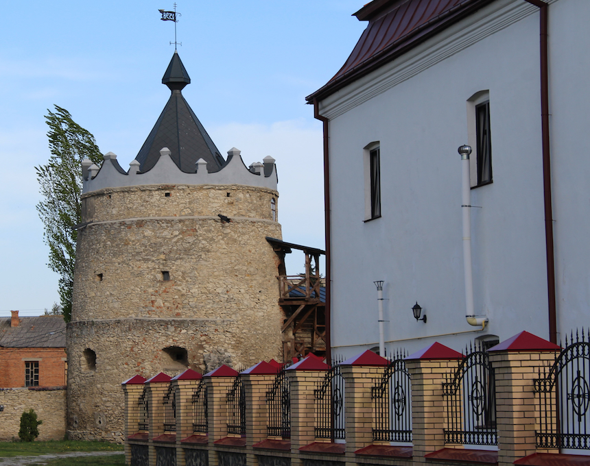 Stone tower of the Latyczów Castle with a conical roof and battlements, adjacent to a white building with red roof accents and decorative fencing.