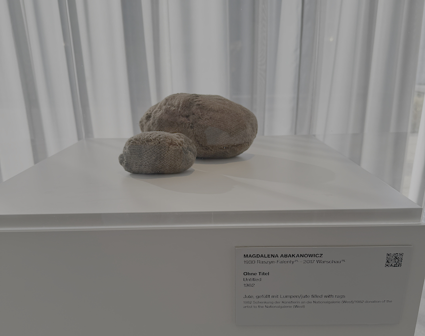 Two irregular, organic-shaped sculptures by Magdalena Abakanowicz from the 'Embriologia' series, made of jute sacks filled with soft material, displayed on a white pedestal in Neue Nationalgalerie, Berlin.