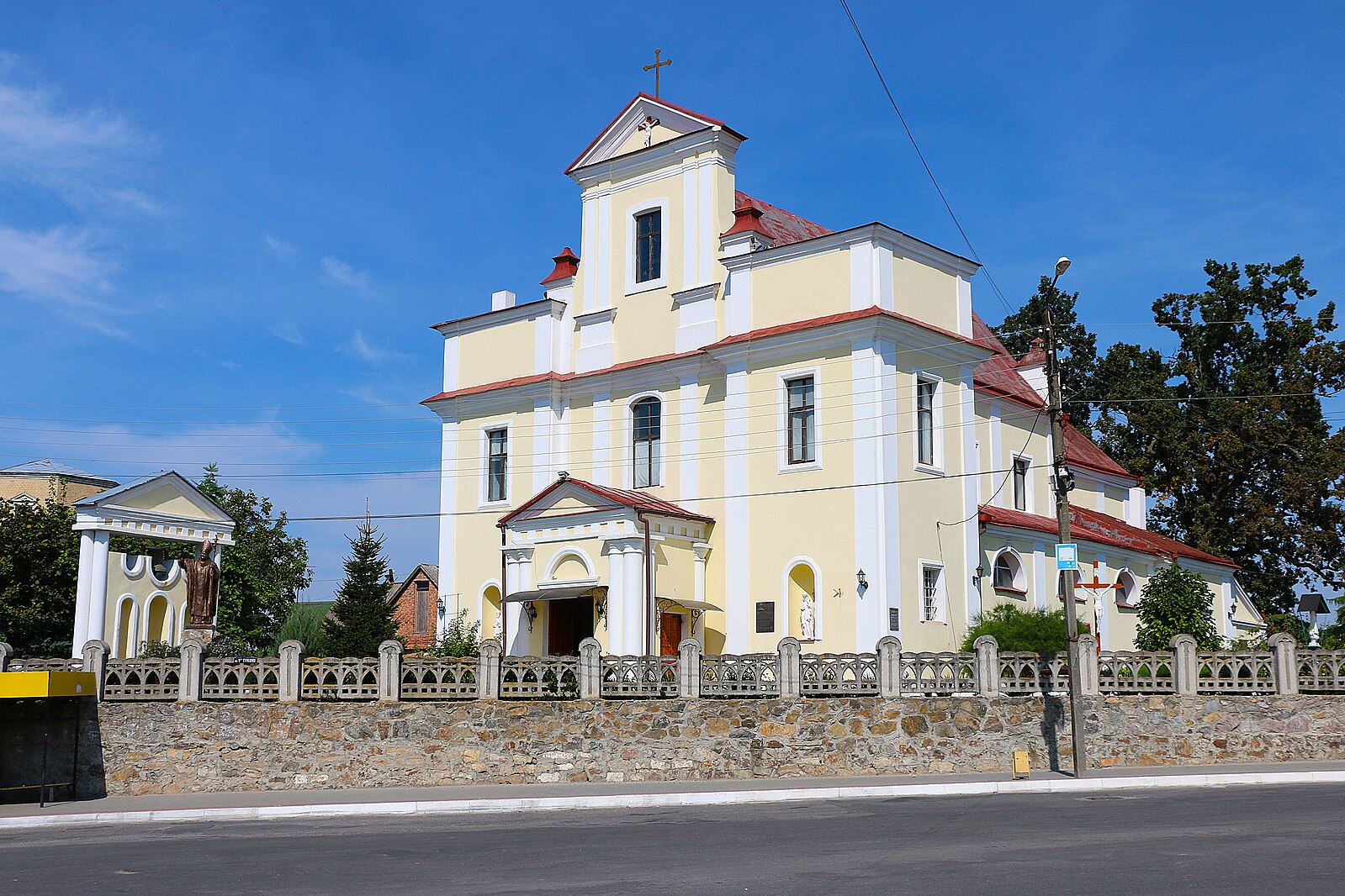 Церква Святої Трійці в Хмільнику, жовто-біла барокова будівля з червоним дахом, оточена кам'яним парканом. На задньому плані видно статую та дерева.