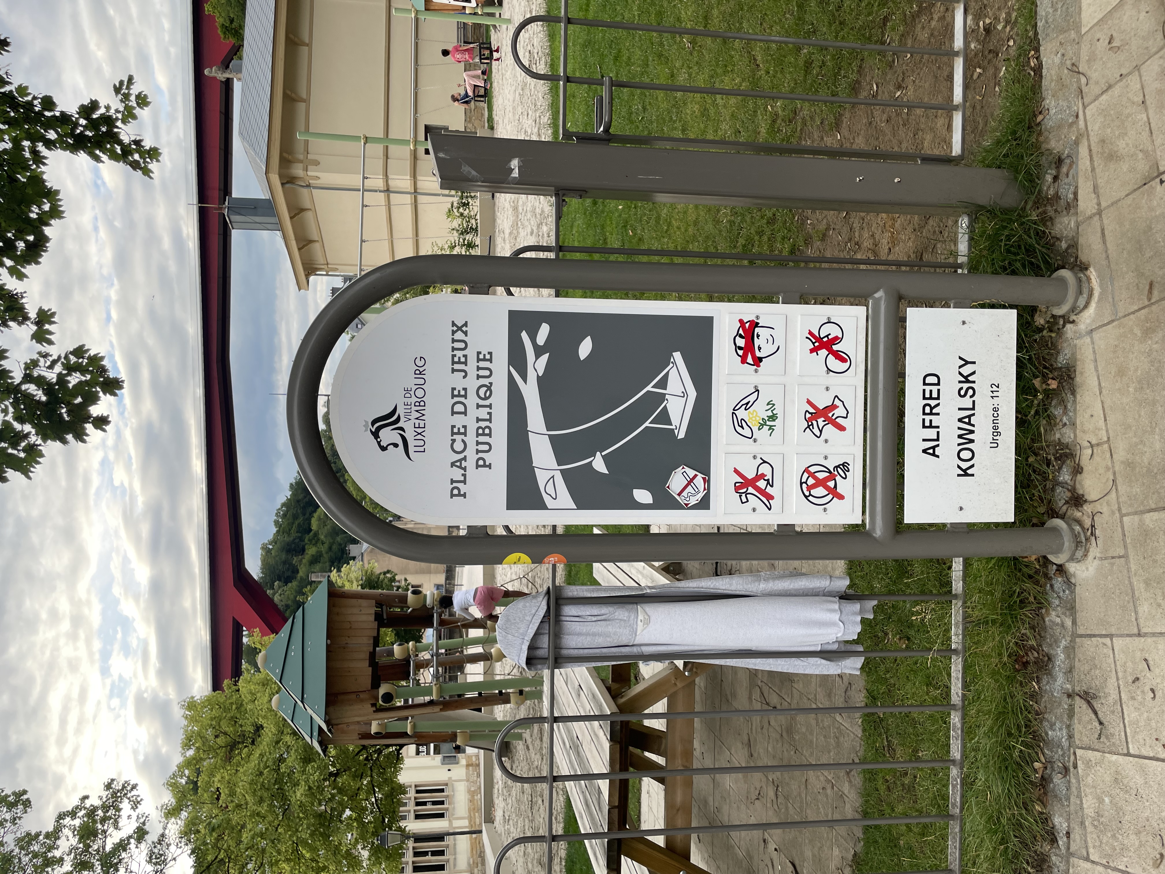 Panneau à l'entrée du terrain de jeux Alfred Kowalsky à Luxembourg, montrant les règles et une structure de jeu en arrière-plan. Un pont rouge et des arbres sont visibles.