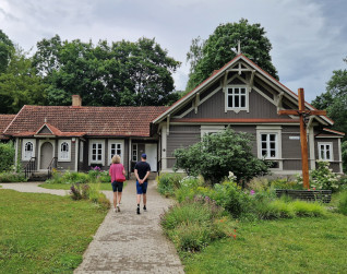 Drewniany dom przy ul. Grybo 29A w Wilnie, gdzie mieszkała św. Faustyna Kowalska. Budynek ma brązową elewację, czerwony dach i krzyż w ogrodzie. Dwie osoby idą ścieżką do wejścia.