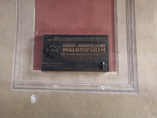 Tablica poświęcona Andrzejowi Małkowskiemu, twórcy harcerstwa polskiego, umieszczona na ścianie z czerwono-beżowym tłem. Tablica zawiera emblemat harcerski i tekst upamiętniający założenie pierwszej drużyny skautowej we Lwowie.